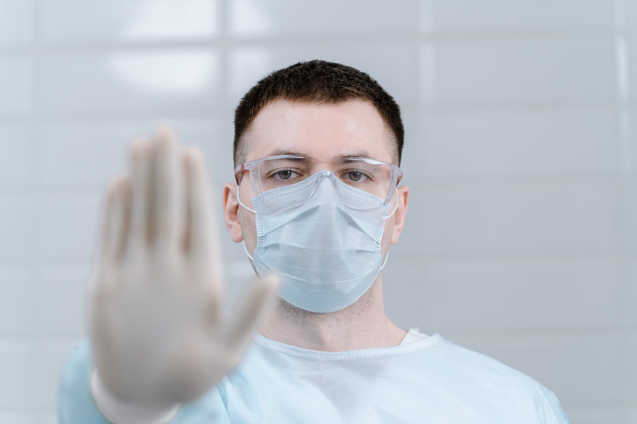 services-01 Healthcare worker wearing protective gear and mask making stop gesture in hospital setting.